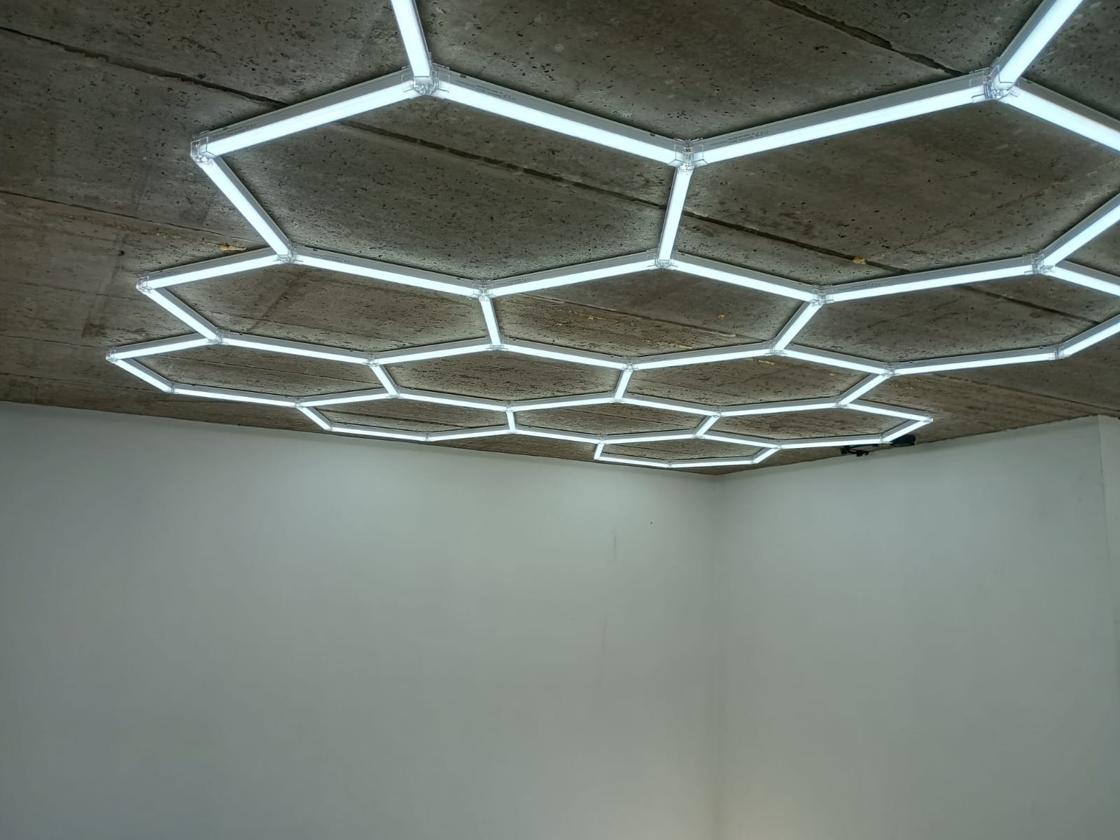 Geometryczna instalacja oświetleniowa LED w kształcie plastra miodu na surowym betonowym suficie. Nowoczesny design, jasne światło. Widok z dołu na białe ściany.