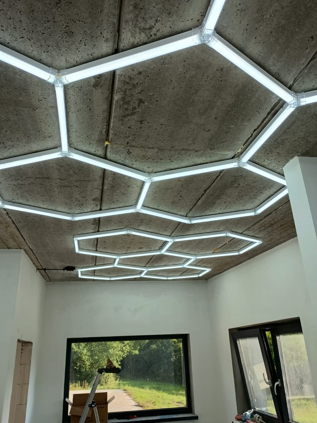 Geometryczna instalacja oświetleniowa LED na surowym betonowym suficie w pomieszczeniu z oknami i drabiną, widok z dołu.