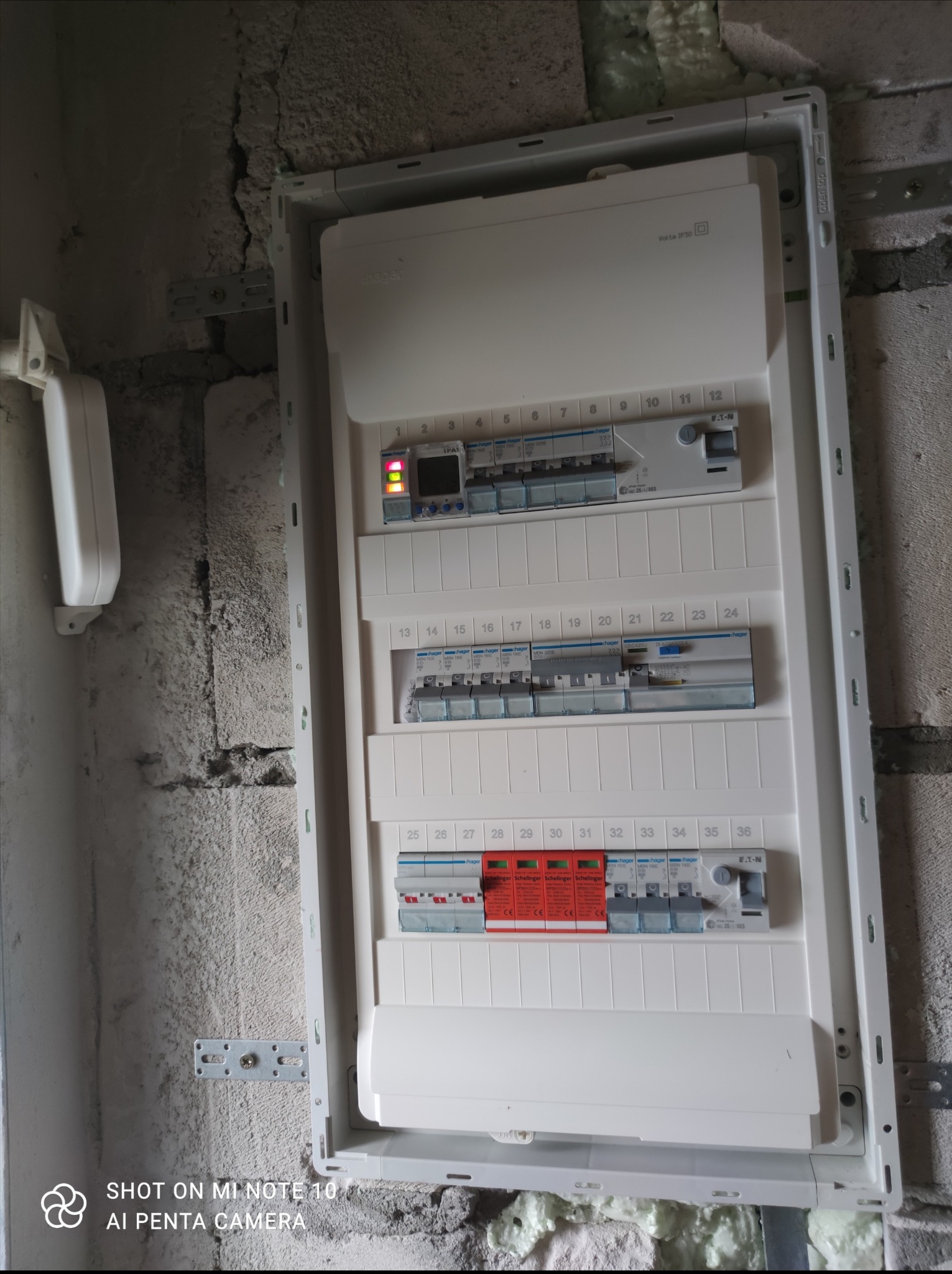 Rozdzielnica elektryczna zamontowana na surowej ścianie, widoczne zabezpieczenia przepięciowe, wyłączniki nadprądowe i programator czasowy, instalacja w trakcie realizacji.