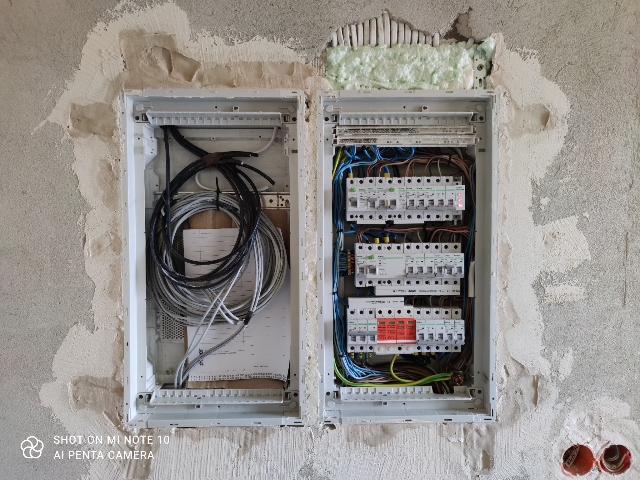 Rozdzielnica elektryczna dwusekcyjna w trakcie montażu w surowej ścianie, widoczne kable, zabezpieczenia i pianka montażowa.