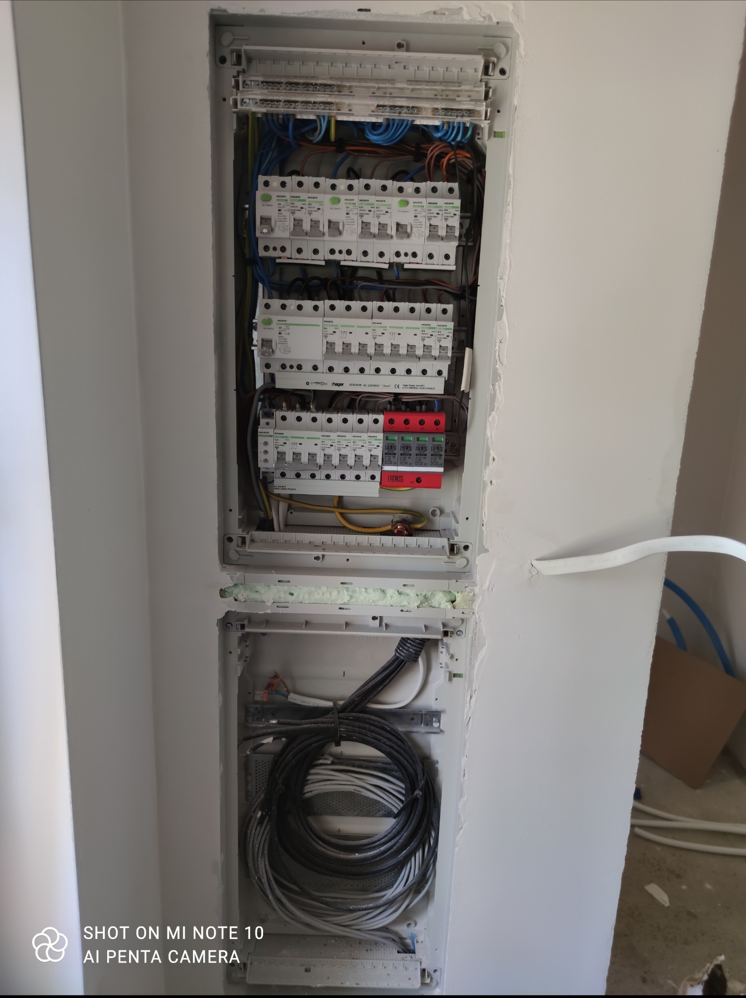 Rozdzielnica elektryczna z zamontowanymi bezpiecznikami, zabezpieczeniami przepięciowymi i okablowaniem, widoczna wnęka w ścianie z pianką montażową i kable w dolnej części.