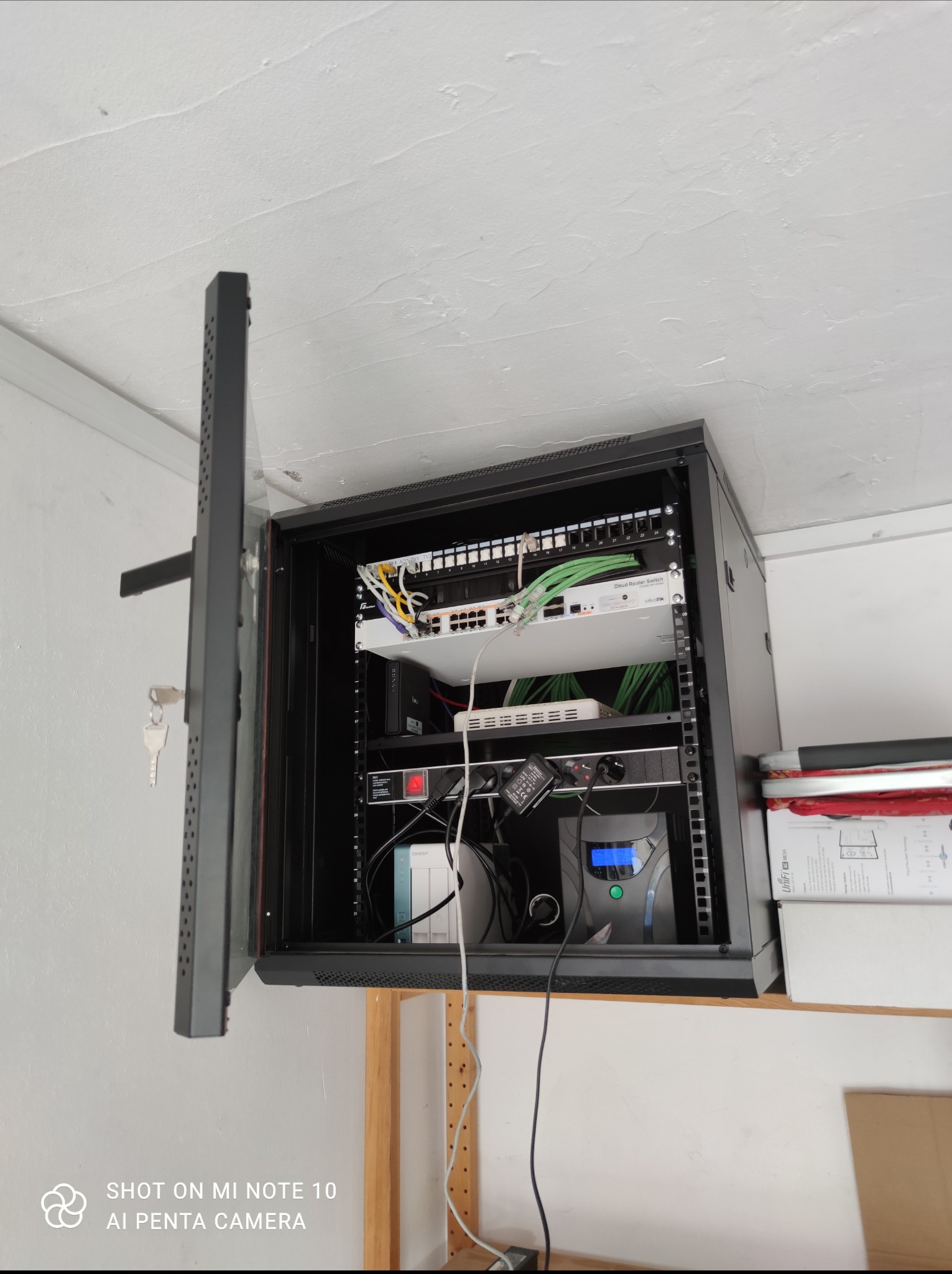 Otwarta szafa rack z zamontowanym sprzętem sieciowym: patch panel, router, switch z zielonymi kablami, UPS z niebieskim wyświetlaczem, na ścianie z widoczną fakturą, klucze w zamku.