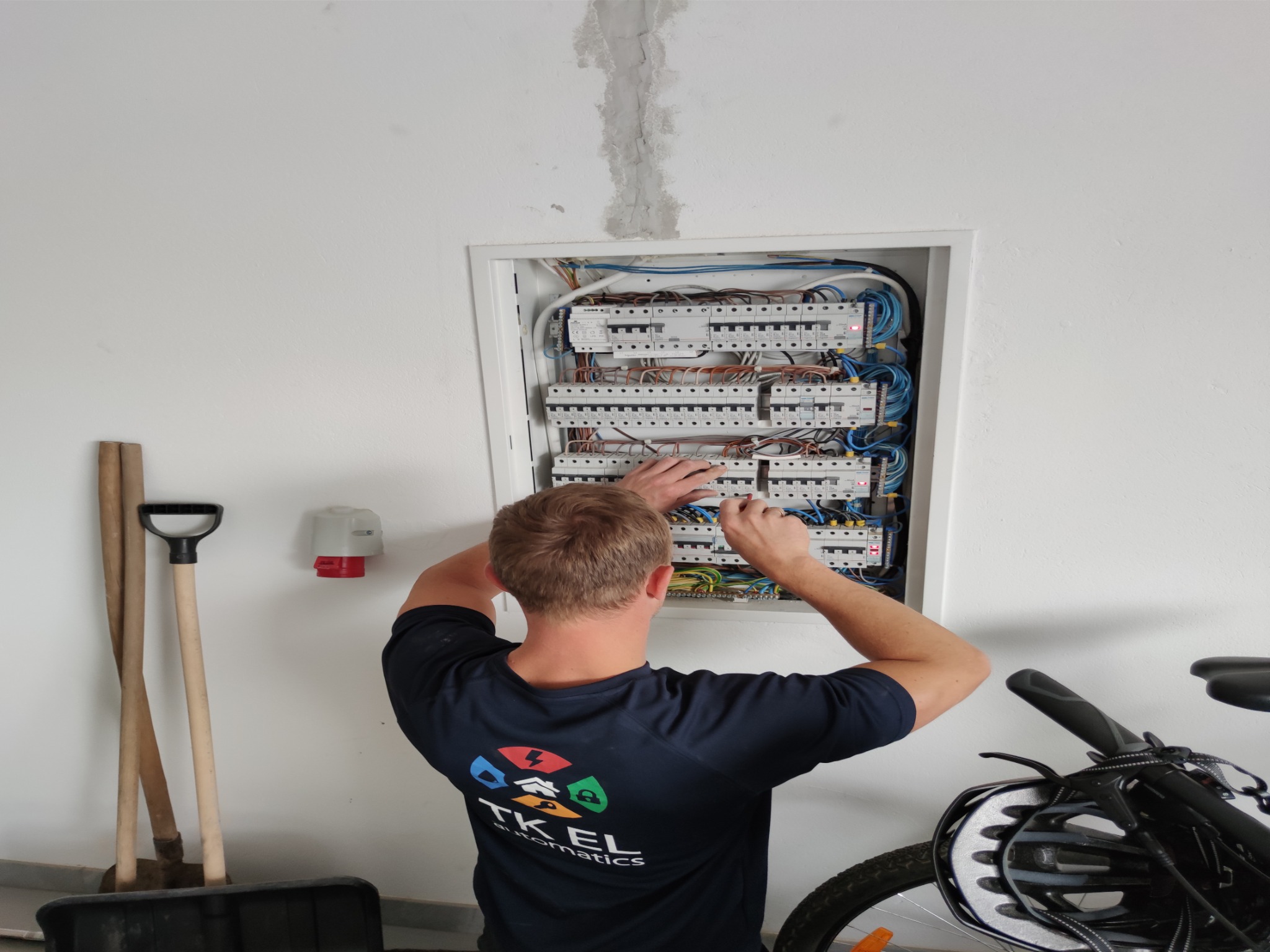 Mężczyzna w koszulce z logo TK EL automatics pracuje przy otwartej skrzynce z bezpiecznikami, widoczne liczne przewody i wyłączniki, obok ściany oparte łopaty i stojący rower.