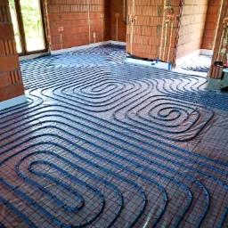 Instalacja ogrzewania podłogowego z niebieskich rur ułożonych spiralnie na czerwonej siatce w surowym wnętrzu budynku z ceglanymi ścianami i widocznymi puszkami elektrycznymi.