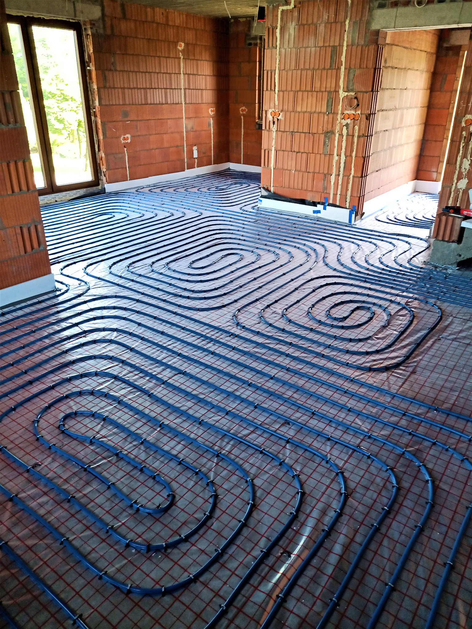 Instalacja ogrzewania podłogowego z niebieskich rur ułożonych spiralnie na czerwonej siatce w surowym wnętrzu budynku z ceglanymi ścianami i widocznymi puszkami elektrycznymi.
