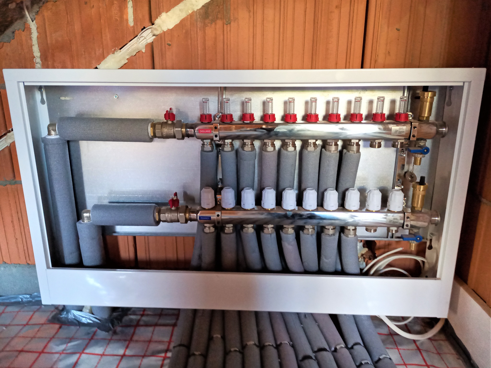 Rozdzielacz ogrzewania podłogowego w białej obudowie, widoczne rury z szarą izolacją i czerwone zawory, instalacja na tle czerwonej cegły i folii z siatką ogrzewania podłogowego.