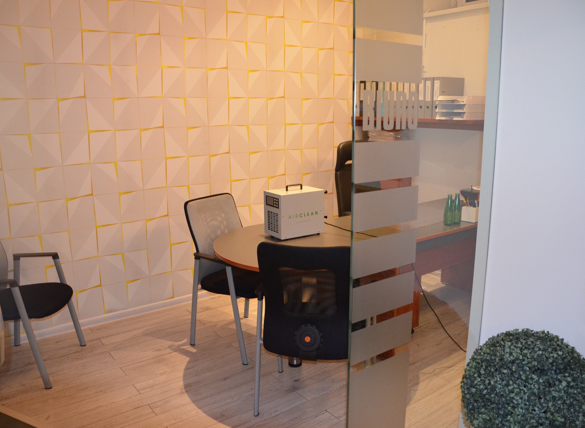 Urządzenie AirClean na okrągłym stole w biurze z geometryczną ścianą, prawdopodobnie używane do dezynfekcji lub oczyszczania powietrza.