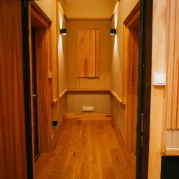 Wnętrze wyciszonego korytarza z drewnianą podłogą, panelem akustycznym na ścianie i dwoma drzwiami z futrynami z drewna, oświetlonym przez dwa kinkiety