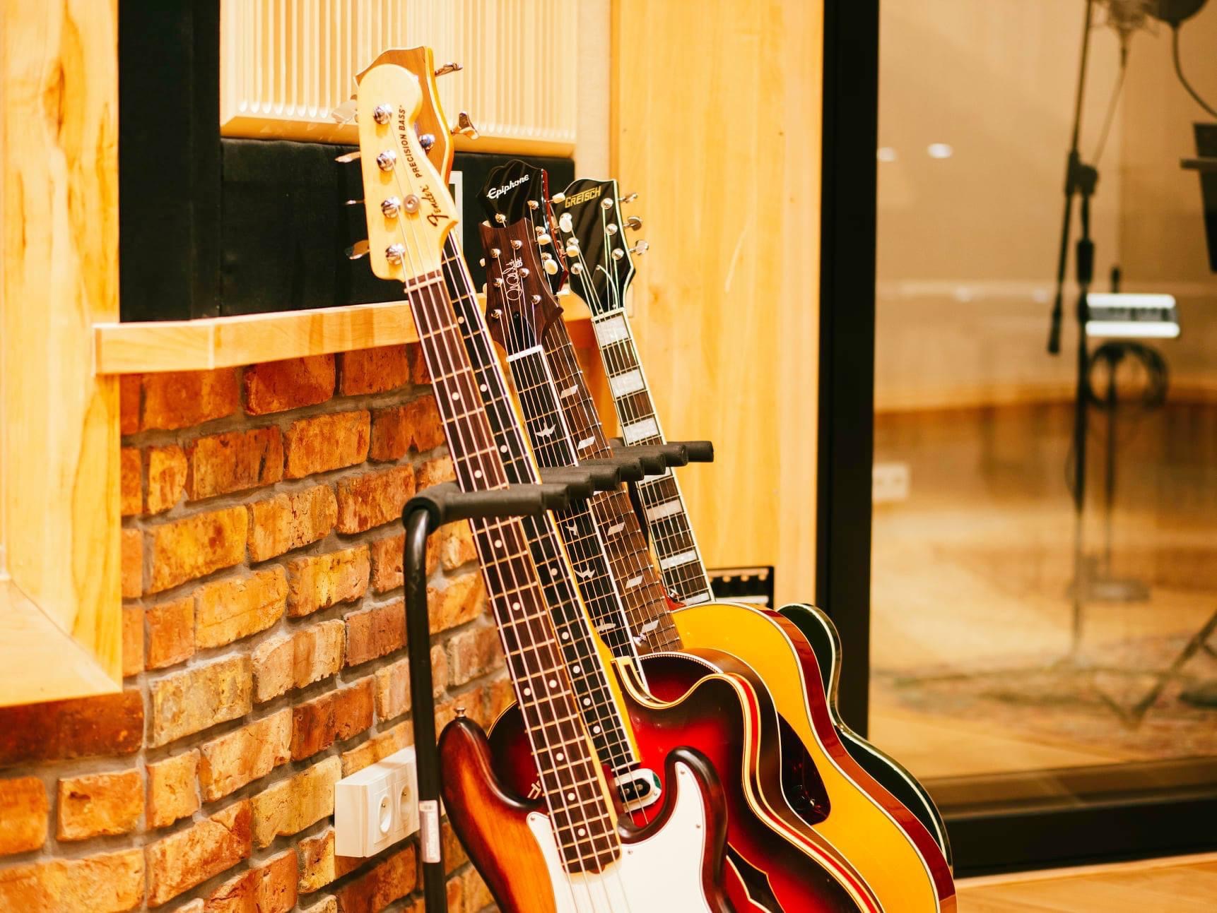 Różne gitary (Fender Precision Bass, Epiphone, Gretsch) ustawione na stojaku przy ceglanej ścianie w pomieszczeniu z mikrofonem studyjnym widocznym w tle.