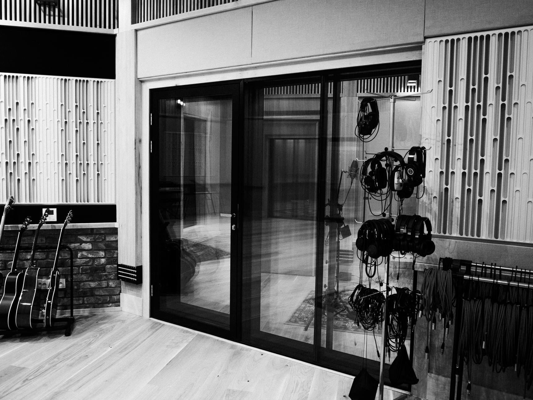 Wnętrze pomieszczenia z akustycznymi panelami ściennymi, trzema gitarami opartymi o ścianę z cegły, przeszklonymi drzwiami przesuwnymi i stojakiem z wieloma parami słuchawek. Widoczne kable...