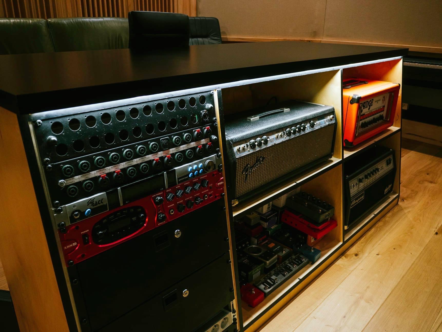 Rząd wzmacniaczy gitarowych i efektów podłączonych do panelu krosowniczego w drewnianej obudowie z czarnym blatem w pomieszczeniu z drewnianą podłogą.