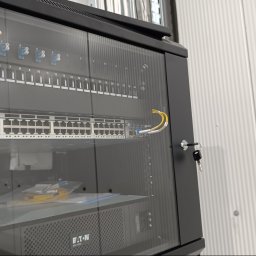 FP SYSTEMS Filip Pawiński - Czarna szafa rackowa z przeszklonymi drzwiami, w których zamocowane są klucze. Wewnątrz widoczny aktywny switch sieciowy z podłączonymi żółtymi kablami światłowodowymi oraz zasilacz UPS Eaton.