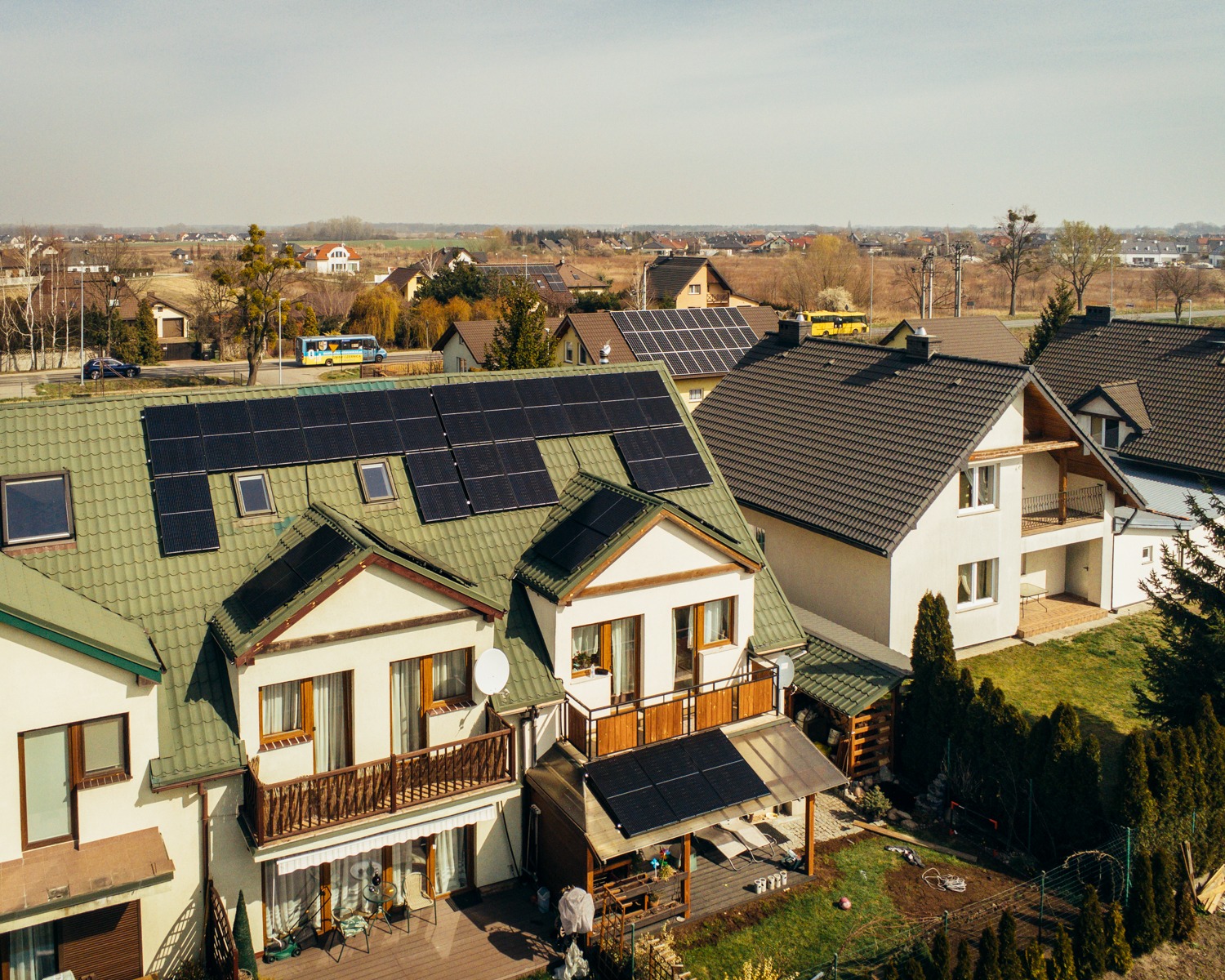 Widok z lotu ptaka na szereg domów jednorodzinnych z panelami słonecznymi na dachach, w tle zabudowa podmiejska i pola uprawne, słoneczny dzień.