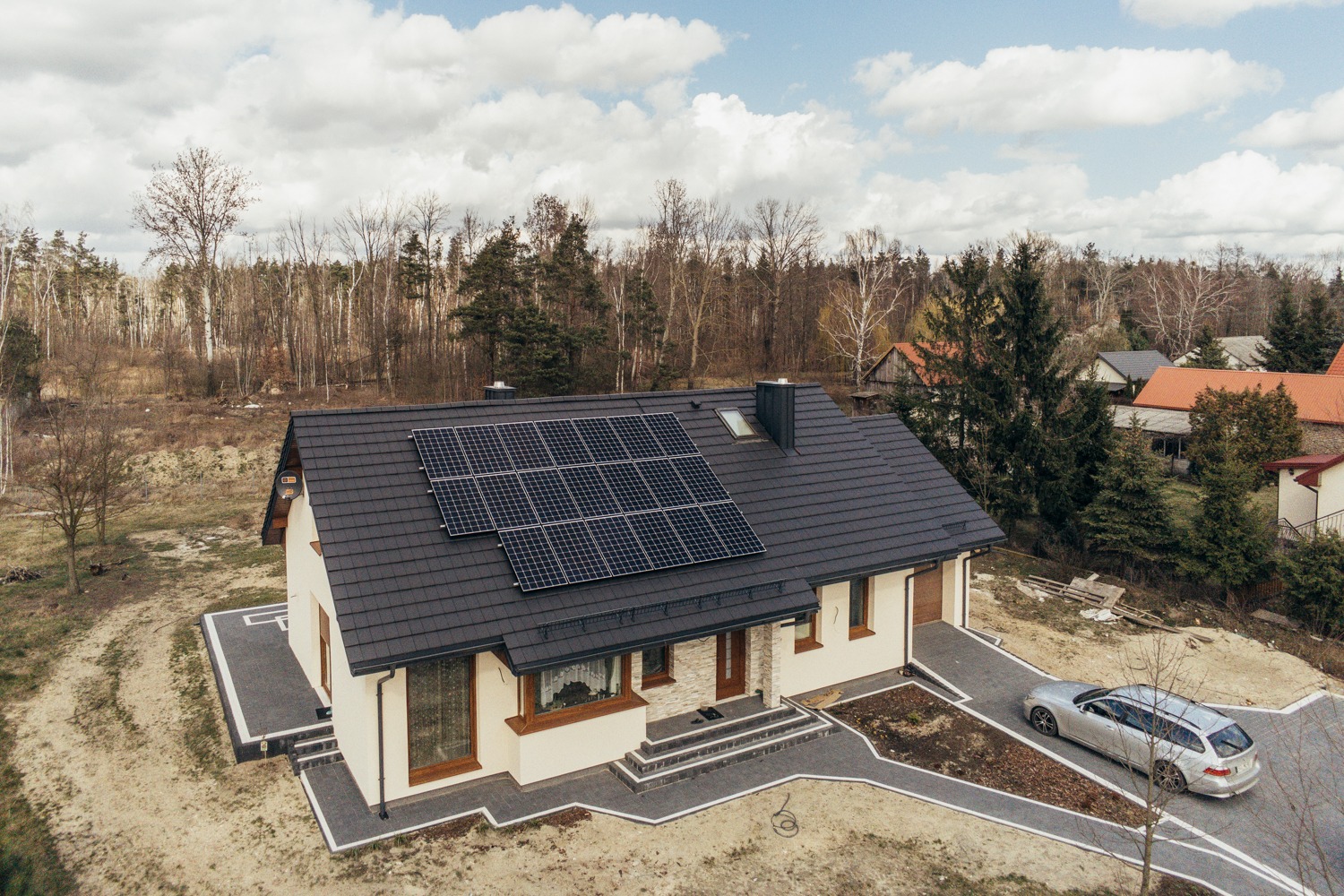 Dom jednorodzinny z panelami słonecznymi na dachu, widok z góry, szara kostka brukowa na podjeździe i samochód kombi w kolorze srebrnym.