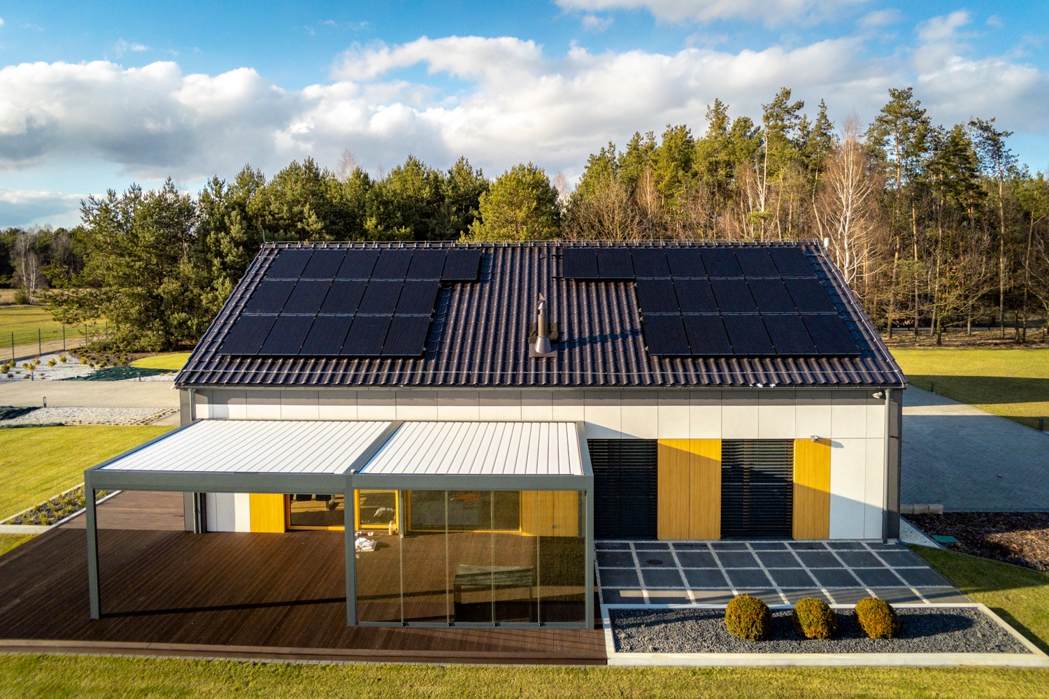 Nowoczesny dom jednorodzinny z panelami słonecznymi na dachu, widok z góry na tle lasu i błękitnego nieba z chmurami.