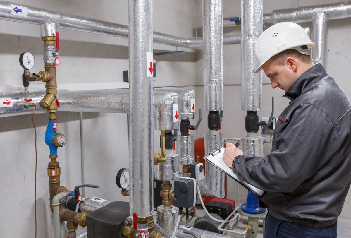 Instalator w kasku, w Trojanowicach, sporządza notatki na tle rur i zaworów instalacji grzewczej, z widocznymi manometrami i izolacją termiczną.