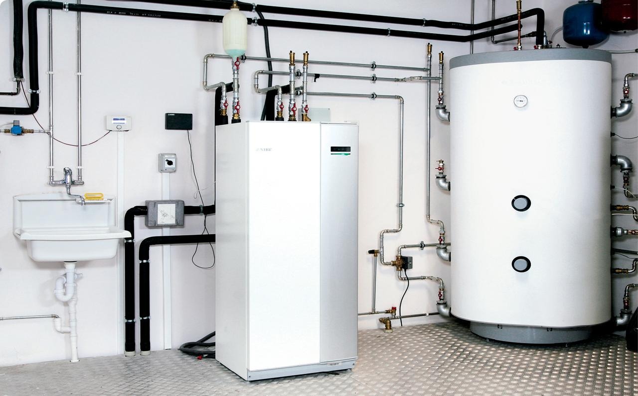 Biała, nowoczesna pompa ciepła NIBE z zbiornikiem wody i instalacją hydrauliczną w kotłowni z metalową podłogą i umywalką