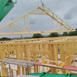 Montaż drewnianej więźby dachowej na konstrukcji domu szkieletowego z użyciem dźwigu, widok na elementy konstrukcyjne i krajobraz w tle.