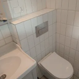 Nowoczesna łazienka z białą ceramiką: umywalka z szafką i wisząca toaleta, białe płytki na ścianach i heksagonalne płytki podłogowe, widoczny fragment gniazdka elektrycznego.