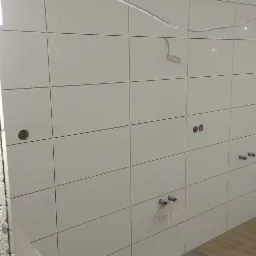 Ściana wyłożona białymi, prostokątnymi płytkami z widocznymi otworami na instalacje wodne i elektryczne, oraz wystającymi przewodami elektrycznymi, podłoga wyłożona panelami imitującymi drewno.