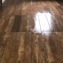 Świeżo położona, błyszcząca podłoga z płytek imitujących drewno, z widocznymi fugami i odbiciami światła, na tle białej ściany w trakcie wykończenia.