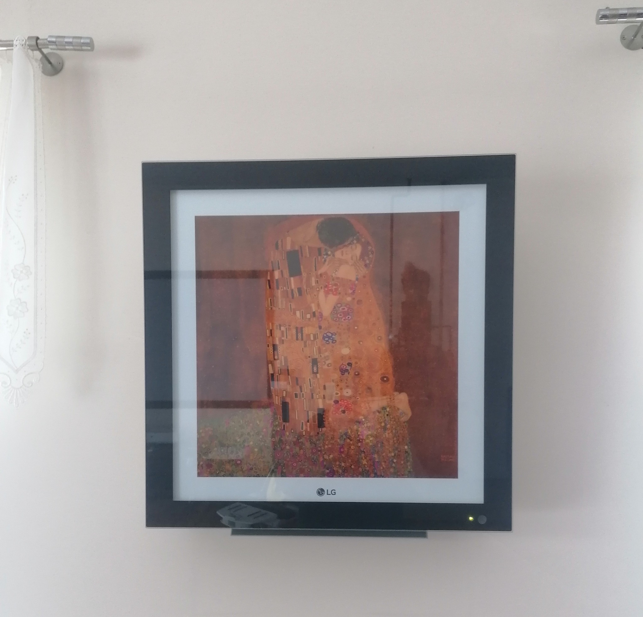Telewizor LG wyświetlający obraz Gustava Klimta 'Pocałunek' w czarnej ramie, zawieszony na jasnej ścianie, obok zasłony.