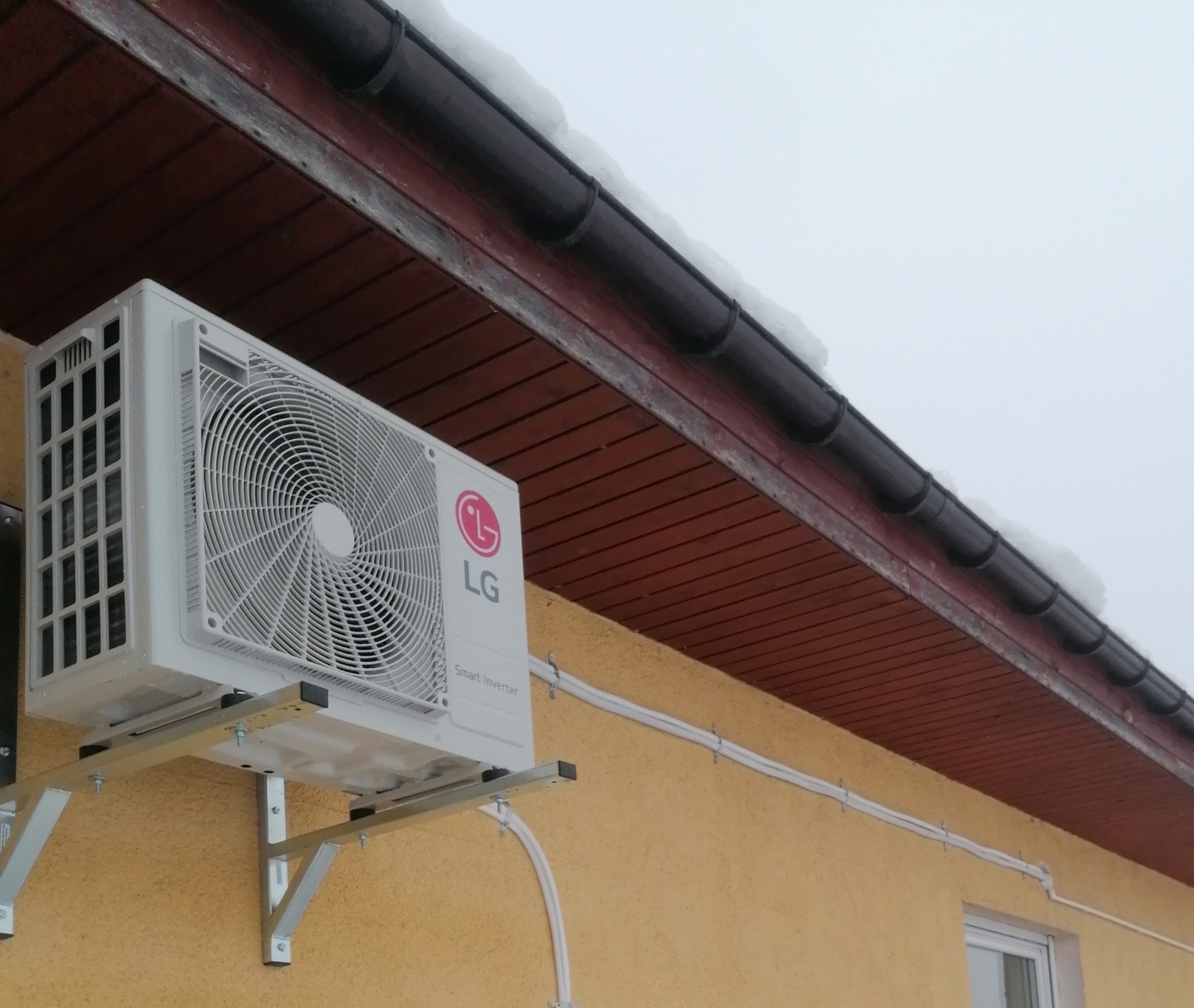 Zewnętrzna jednostka klimatyzacji LG Smart Inverter zamontowana na ścianie budynku z widocznym orynnowaniem i śniegiem na dachu.
