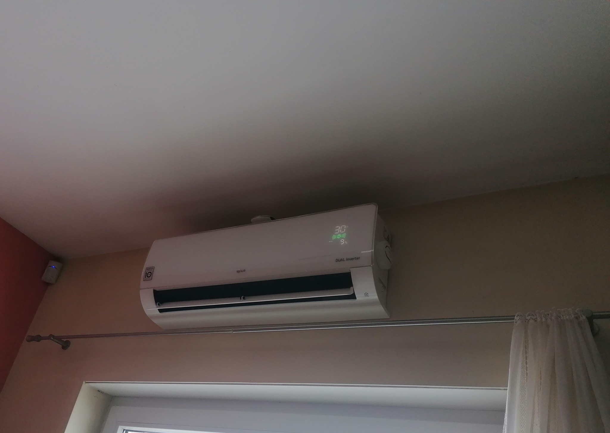 Biała jednostka wewnętrzna klimatyzacji LG Dual Inverter zamontowana na ścianie nad oknem, z widocznym wyświetlaczem temperatury 30 stopni Celsjusza, obok karnisz z zasłoną.