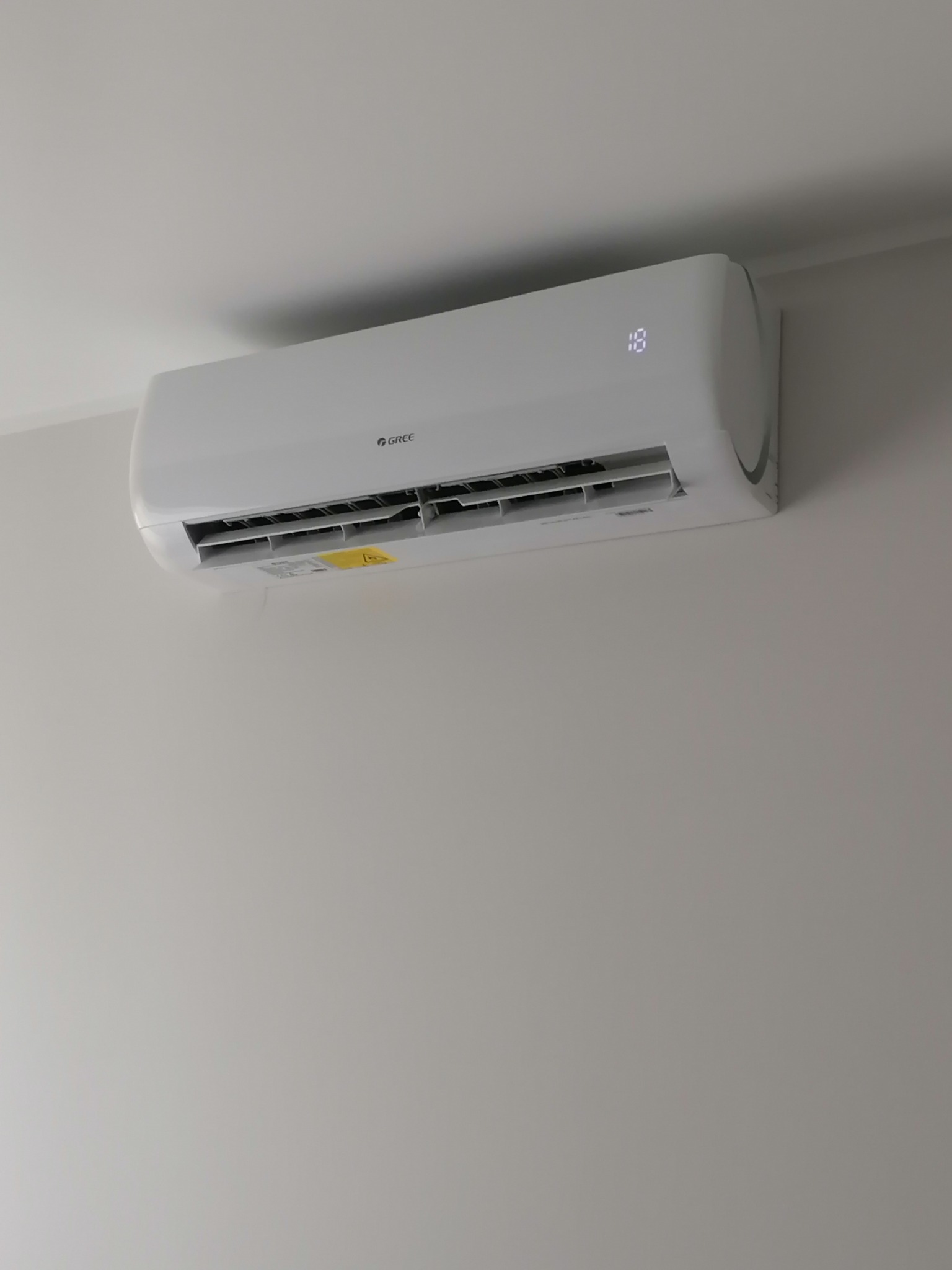 Biała jednostka klimatyzacji GREE MUSE, zamontowana na jasnej ścianie, wyświetlająca temperaturę 18 stopni Celsjusza.
