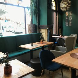 Karpińscy Firma Budowlana - Wnętrze kawiarni z zielonymi ścianami, drewnianymi stolikami, pluszowymi krzesłami i kanapą, dekoracyjną lampą oraz roślinami doniczkowymi, widok na ulicę przez okno.