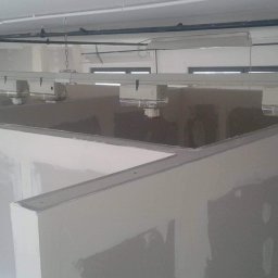 Karpińscy Firma Budowlana - Wnętrze w trakcie prac wykończeniowych: surowe ściany z płyt gipsowo-kartonowych, widoczne łączenia wymagające szpachlowania, oświetlenie przemysłowe podwieszone na łańcuchach, fragment okna w tle.