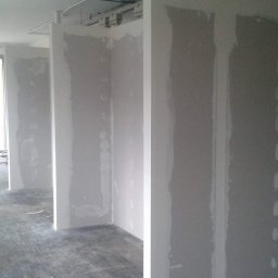 Karpińscy Firma Budowlana - Montaż ścian działowych z płyt gipsowo-kartonowych w surowym wnętrzu biurowym, widoczne szpachlowane łączenia, surowy beton na podłodze, instalacje pod sufitem.