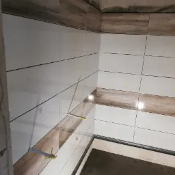 Ściana łazienki w trakcie układania płytek: białe płytki z drewnopodobnym pasem, widoczne szyny poziomujące i fragment surowego betonu.