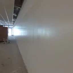 Świeżo pomalowana, jasna ściana w biurze, widoczna instalacja podwieszanego sufitu i niedokończona podłoga, perspektywa z niskiego kąta.