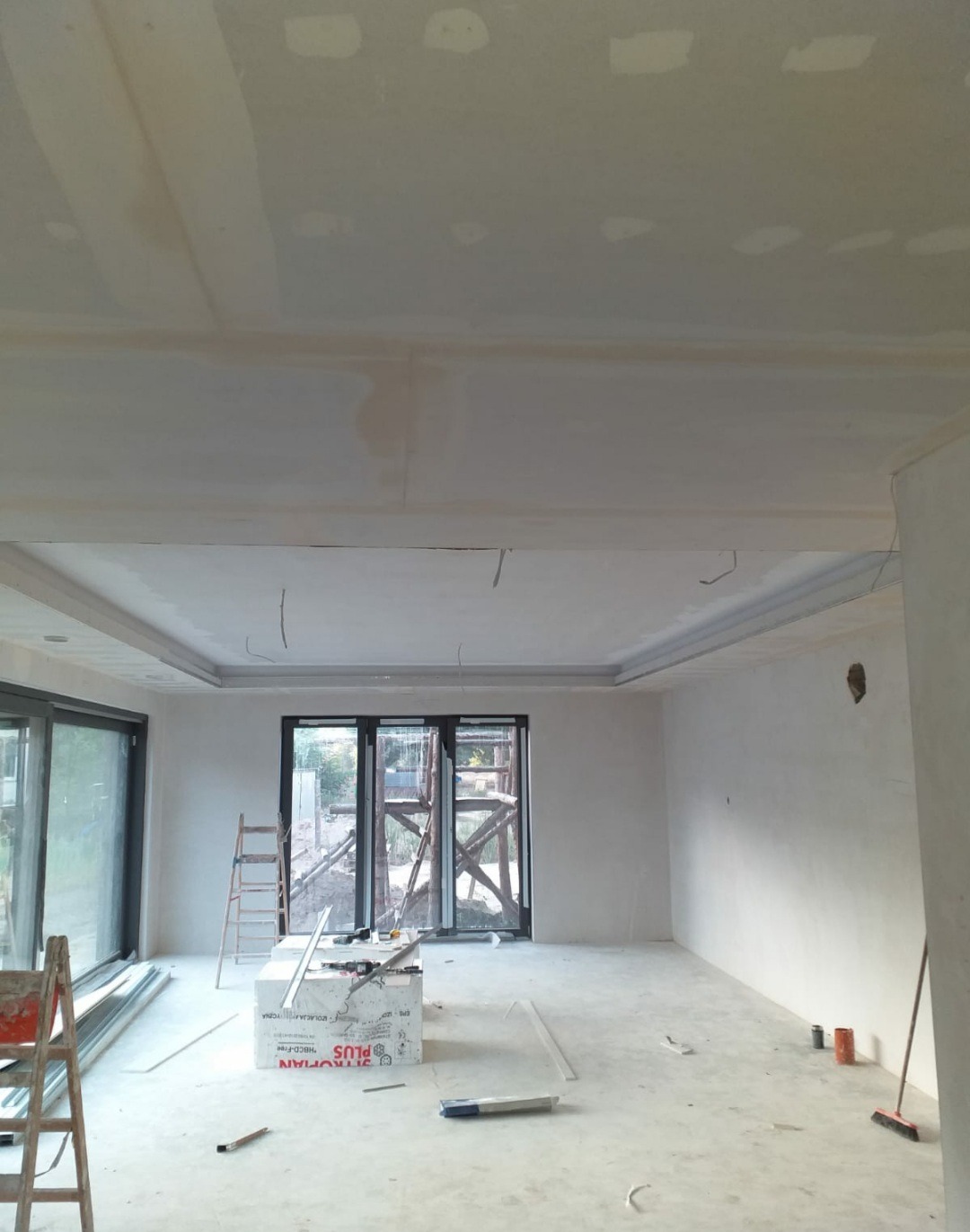 Wnętrze w trakcie prac wykończeniowych: sufit podwieszany z płyt gipsowo-kartonowych, szpachlowane łączenia, widoczne okablowanie, drabiny, materiały budowlane i narzędzia na podłodze, duże okna...