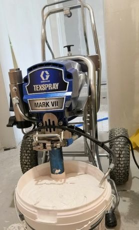 Profesjonalny agregat malarski Graco TexSpray Mark VII zanurzony w wiadrze z farbą w trakcie przygotowań do malowania natryskowego ścian w pomieszczeniu.