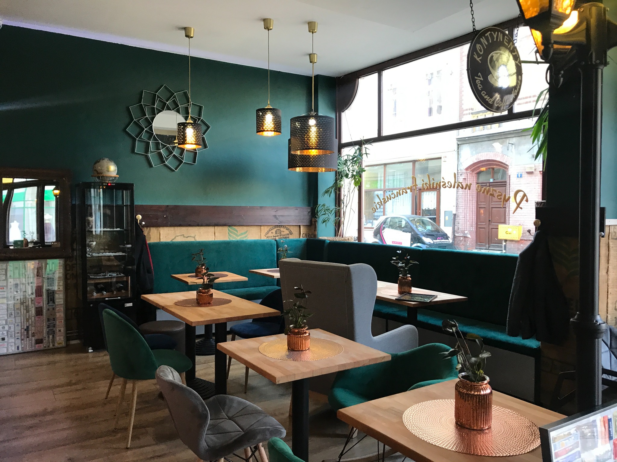 Wnętrze kawiarni z ciemnozielonymi ścianami, drewnianymi stołami z miedzianymi podkładkami i roślinami w doniczkach, oświetlone wiszącymi lampami z ażurowymi kloszami.