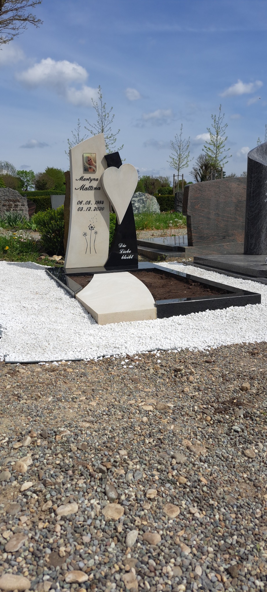 Jasny nagrobek z fotografią i sercem, otoczony białym żwirem, na tle innych nagrobków i zieleni. Kontrastowe kolory i nowoczesny design.