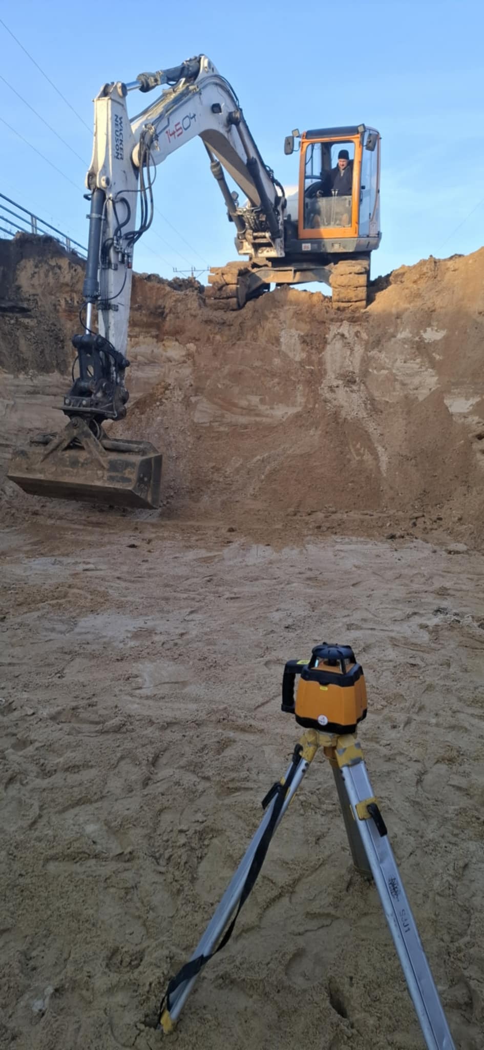 Żółty niwelator laserowy na statywie ustawiony na piaszczystym terenie budowy, w tle koparka Wacker Neuson i wykop ziemny.