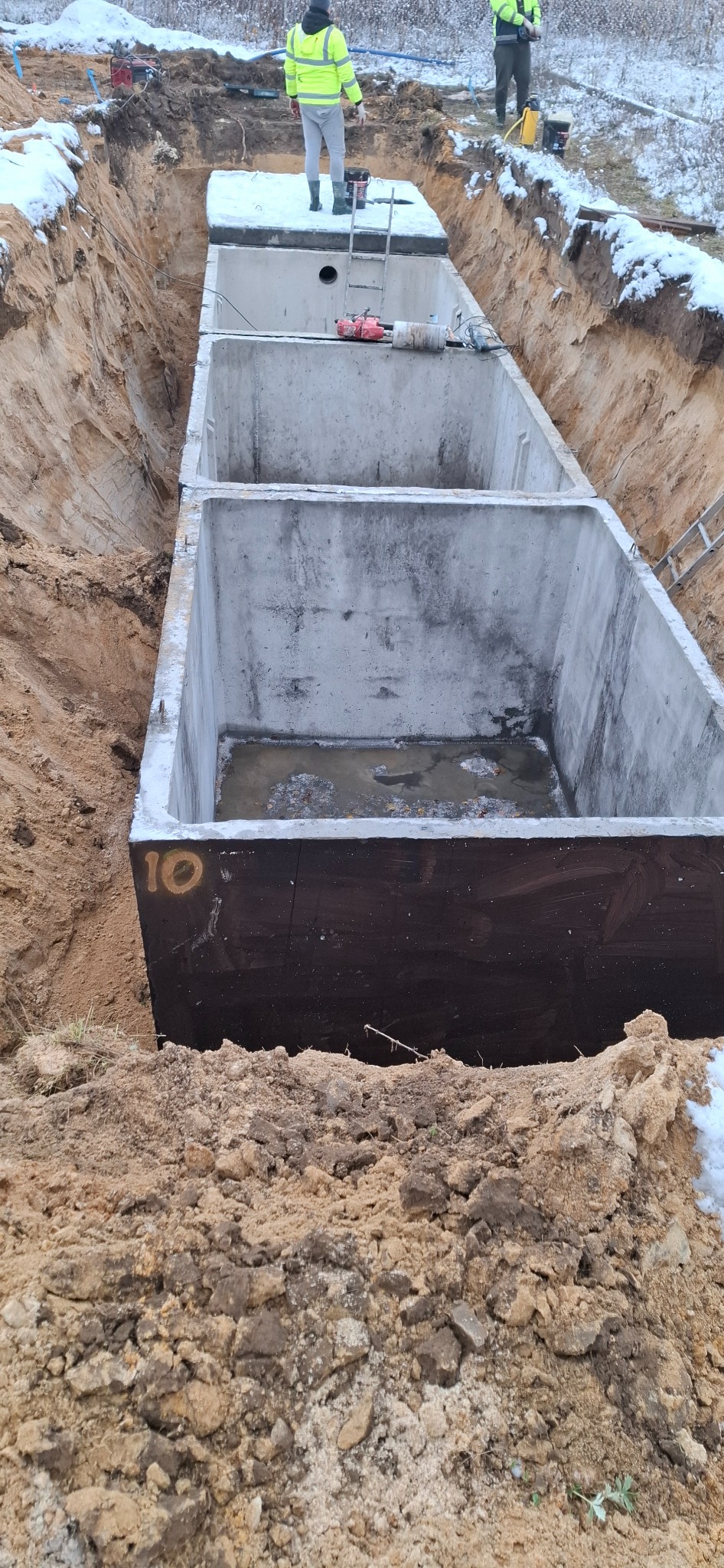 Betonowy zbiornik podzielony na trzy komory, umieszczony w wykopie ziemnym, widoczne osoby w odblaskowych kamizelkach oraz numer '10' na jednej ze ścian zbiornika.