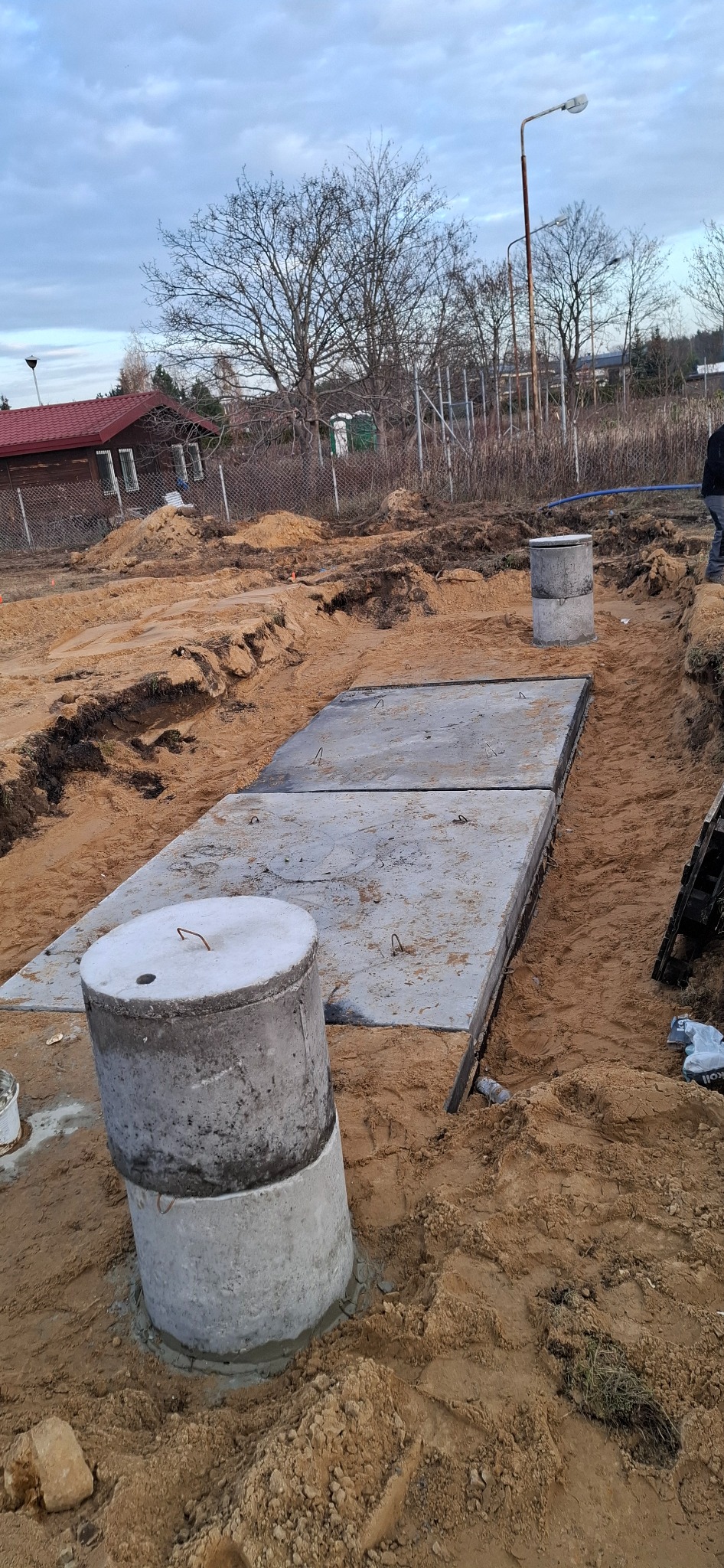 Betonowe elementy przydomowej oczyszczalni ścieków w wykopie, widoczne płyty i studzienki, teren budowy w piasku, pochmurne niebo.