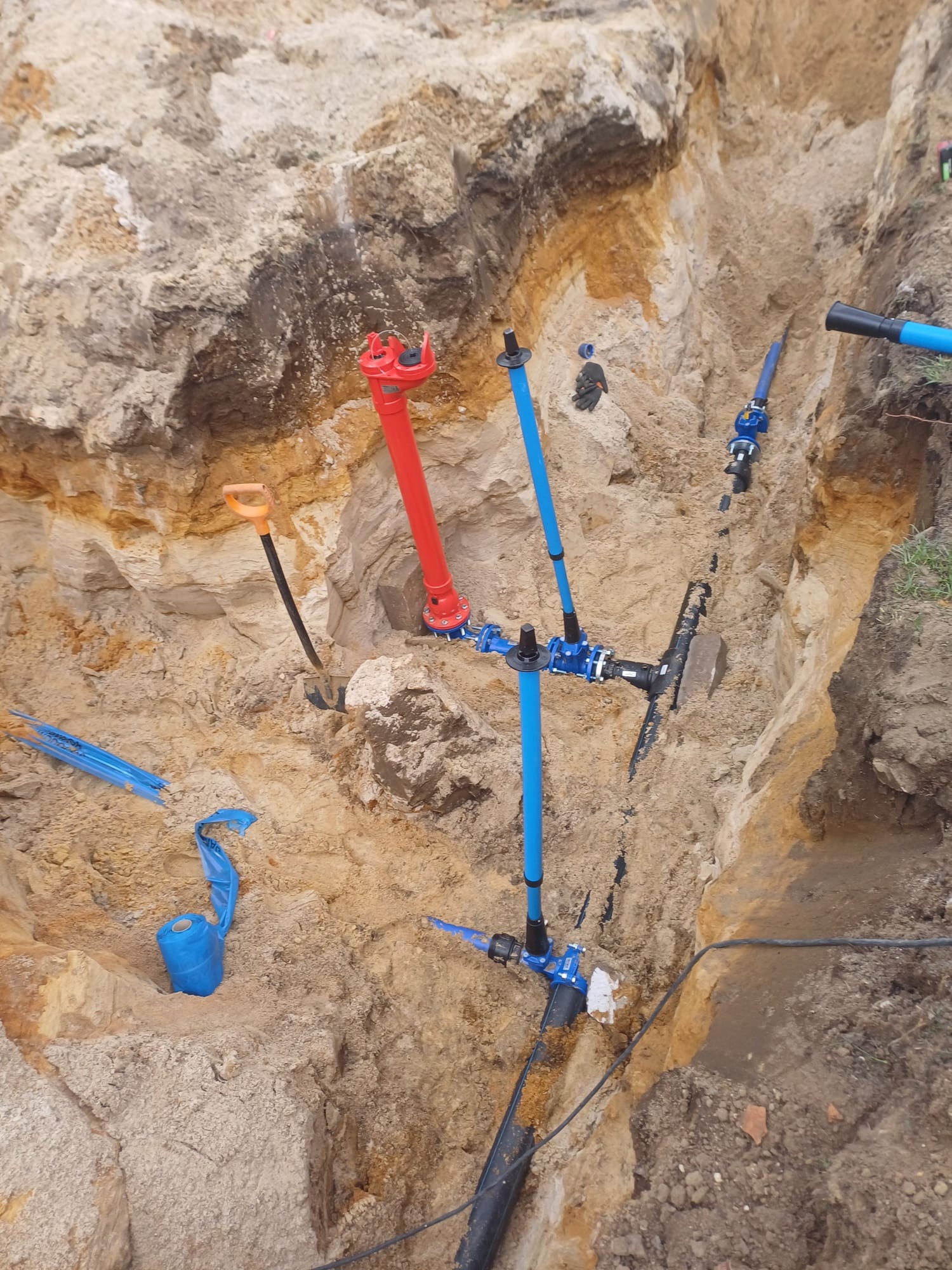 Wykop z widocznymi rurami wodociągowymi w kolorze niebieskim i czerwonym, złączkami i czarną rurą, widoczne narzędzia: szpadel i łopata, ziemia o różnej strukturze i kolorach.