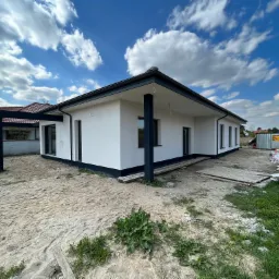 Nowoczesny, parterowy dom z płaskim dachem, białą elewacją i czarnymi ramami okiennymi, widoczny w surowym otoczeniu placu budowy z piaskiem i materiałami budowlanymi.