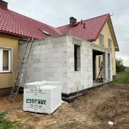 Budowa domu jednorodzinnego w trakcie realizacji, widoczne białe bloczki betonu komórkowego, drabina oparta o ścianę, paleta bloczków marki Prefbet, czerwony dach budynku w tle.