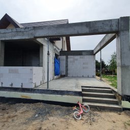 Adrian Zalewski - Surowa konstrukcja tarasu z betonu i bloczków, widoczne zbrojenia i stopnie, na ziemi leży różowy rowerek biegowy.