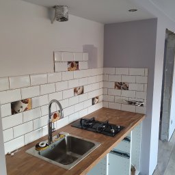 Adrian Zalewski - Montaż płytek ściennych z motywem kawy nad blatem kuchennym z zamontowanym zlewem i płytą gazową, widoczne listwy dystansowe między płytkami, instalacja wentylacyjna w suficie, surowy stan...