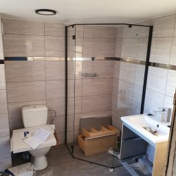 Adrian Zalewski - Remont łazienki: montaż kabiny prysznicowej z czarnymi profilami i umywalki z szafką w trakcie instalacji, widoczne narzędzia i materiały budowlane, płytki imitujące kamień na ścianach.