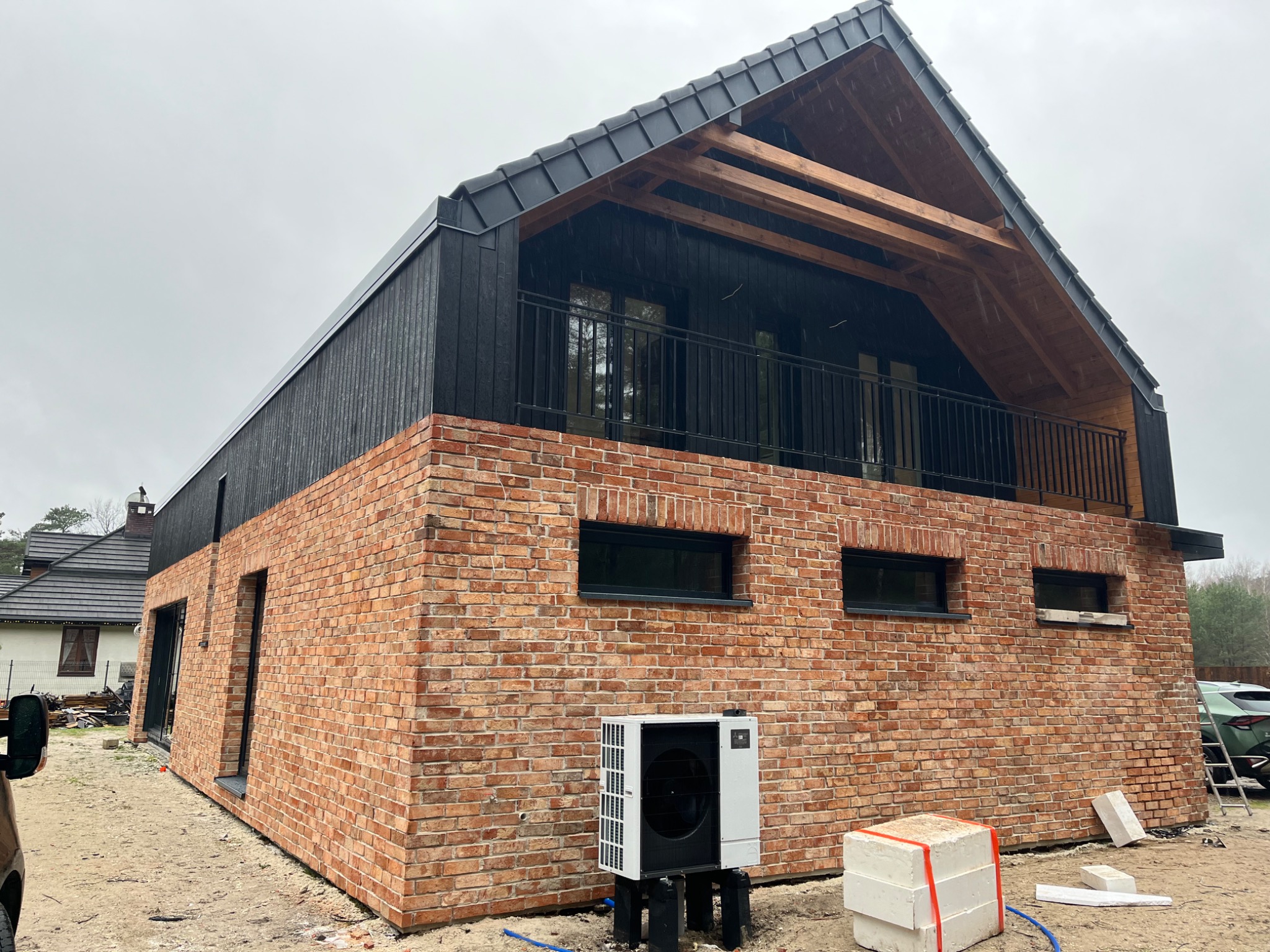 Dwukondygnacyjny dom w trakcie budowy, z czerwoną ceglaną elewacją i czarną drewnianą górną częścią, balkon z czarną balustradą, pompa ciepła na zewnątrz, widoczne materiały budowlane.