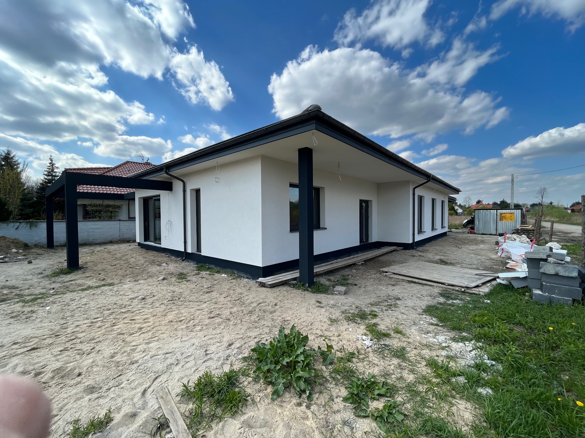 Nowoczesny, parterowy dom z płaskim dachem, białą elewacją i czarnymi ramami okiennymi, widoczny w surowym otoczeniu placu budowy z piaskiem i materiałami budowlanymi.