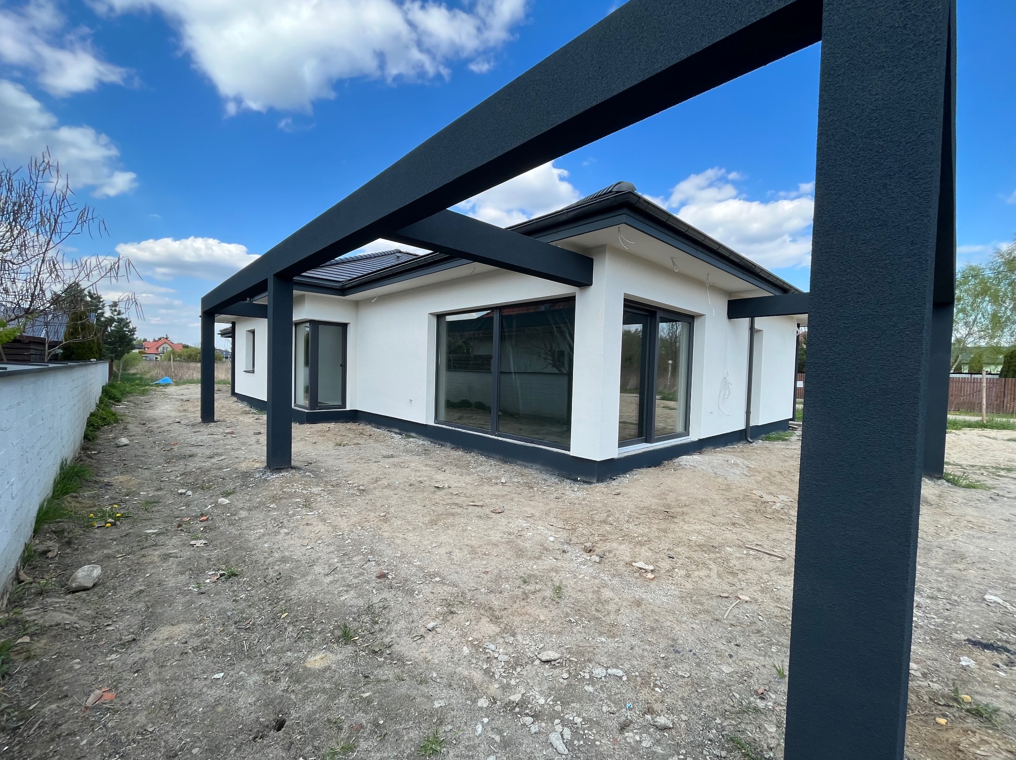 Nowoczesny, parterowy dom z białymi ścianami i dużymi oknami, z elementami konstrukcji tarasu z ciemnymi, grafitowymi słupami i belkami, na działce w trakcie zagospodarowywania.